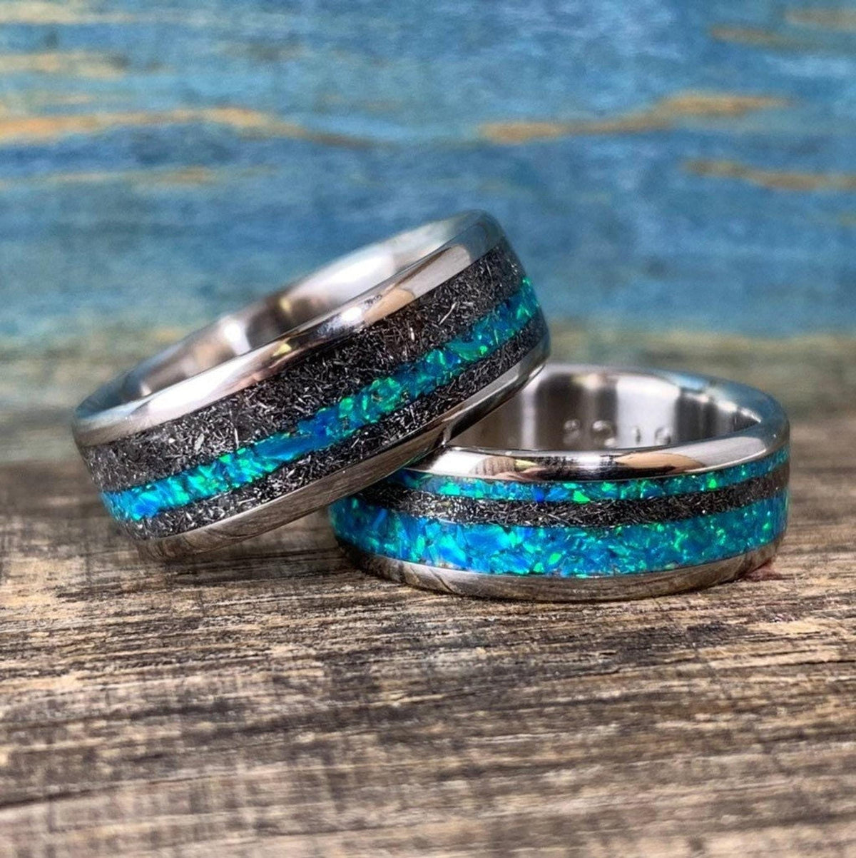 Two wedding bands made of titanium with meteorite inlays and bright blue-green opal stripes rest on a wooden surface.
