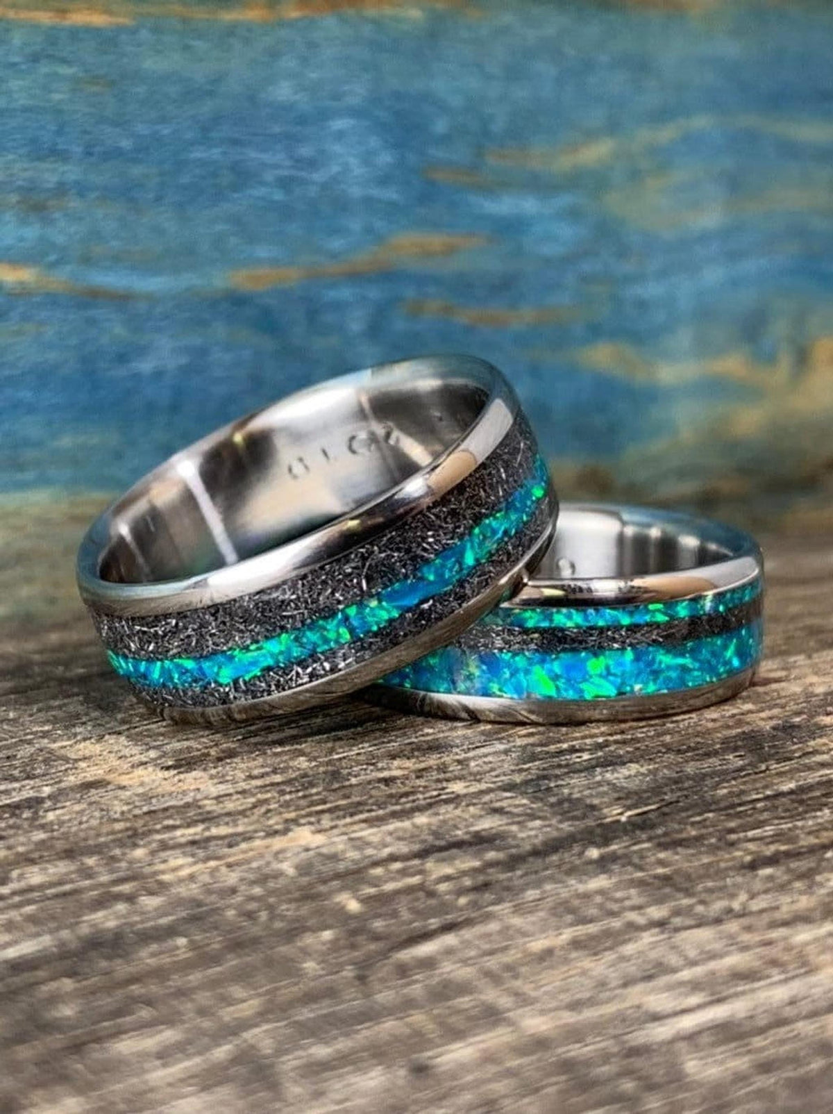 Two titanium wedding rings featuring meteorite bands and central vibrant blue-green opal stripes placed on a rustic wood plank.