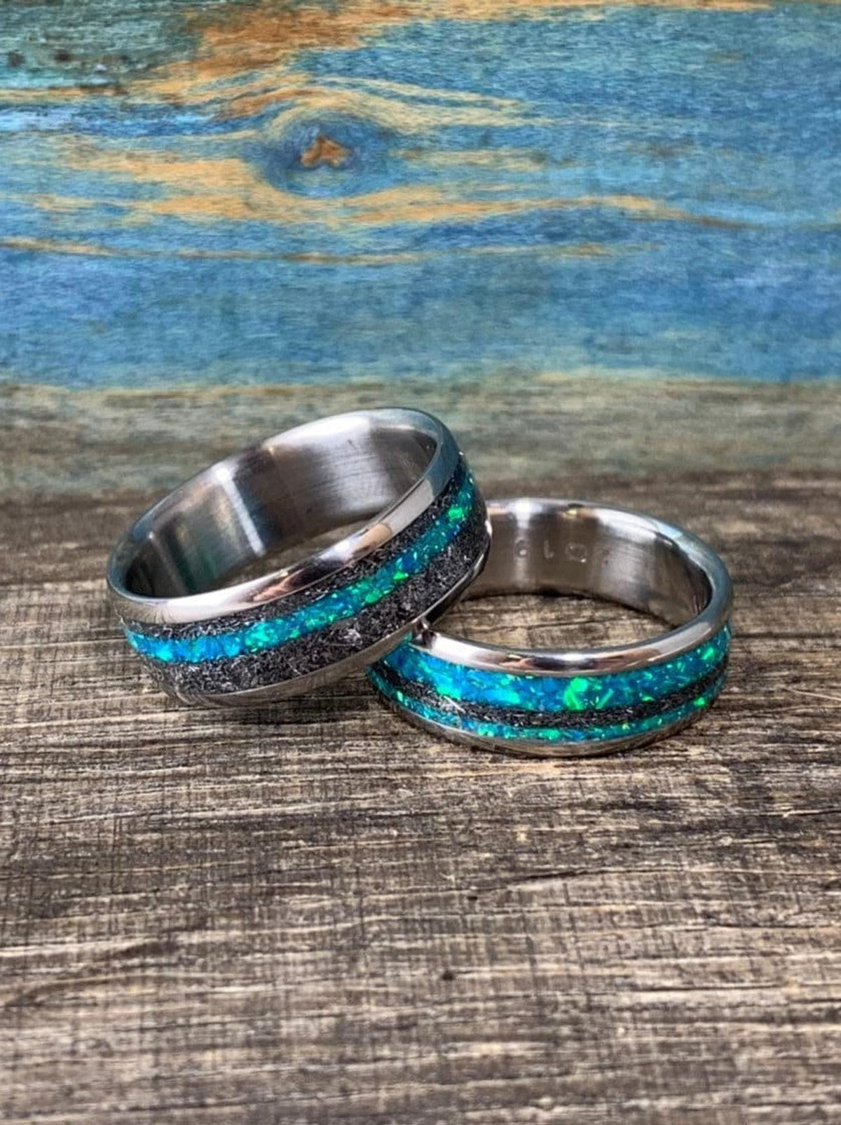 Two titanium rings featuring blue-green opal and textured meteorite inlays displayed on a rustic wooden table.