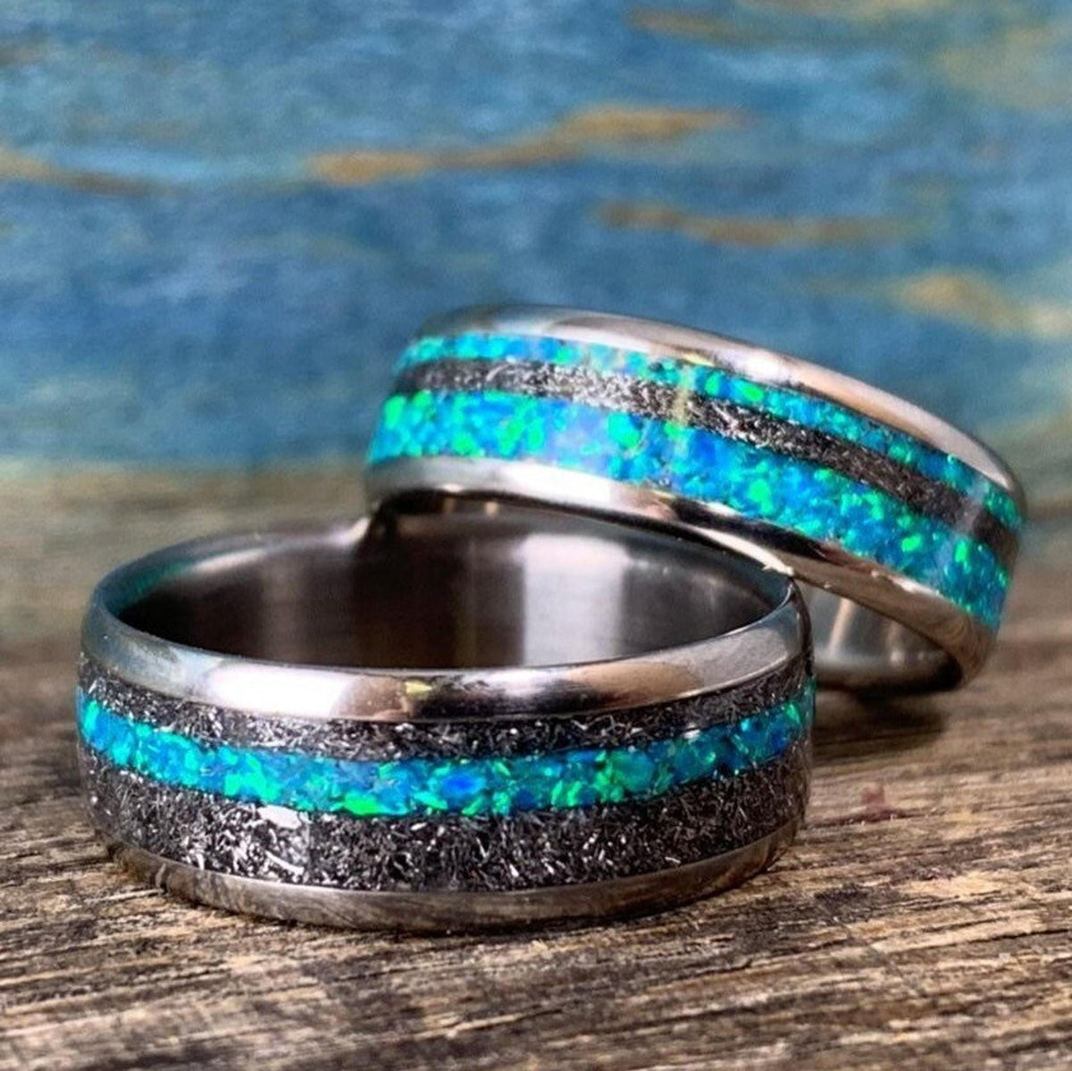 Two titanium wedding bands with meteorite and blue-green opal inlays positioned with one ring lying flat and the other standing behind it.