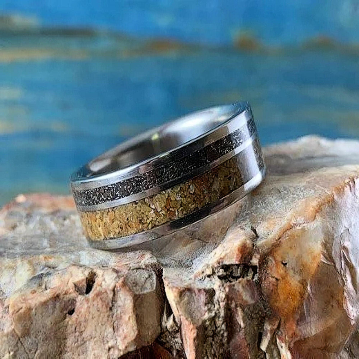 A metal ring with wood and meteorite inlays rests on a rough, textured rock surface.