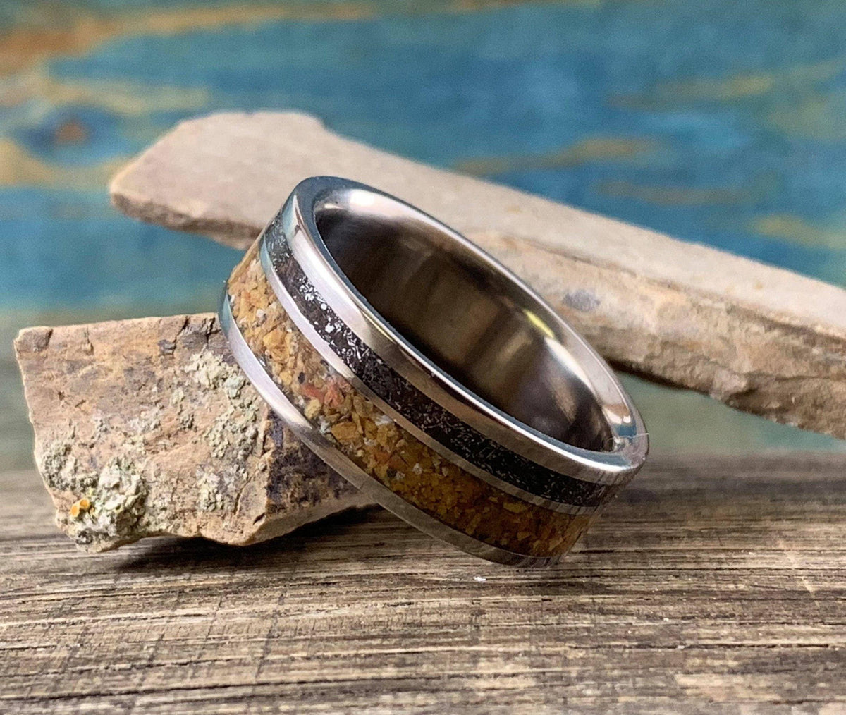A metal ring featuring inlays of textured wood and dark meteorite rests on a weathered wooden board.