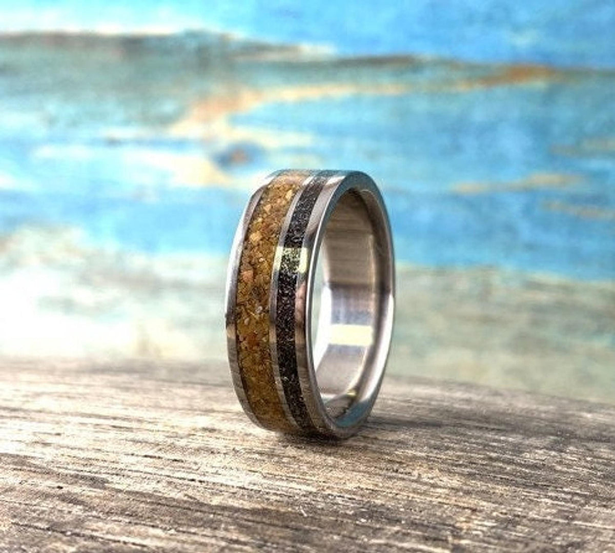 A metal ring featuring a textured wood inlay and a dark meteorite stripe stands upright on a wooden board.