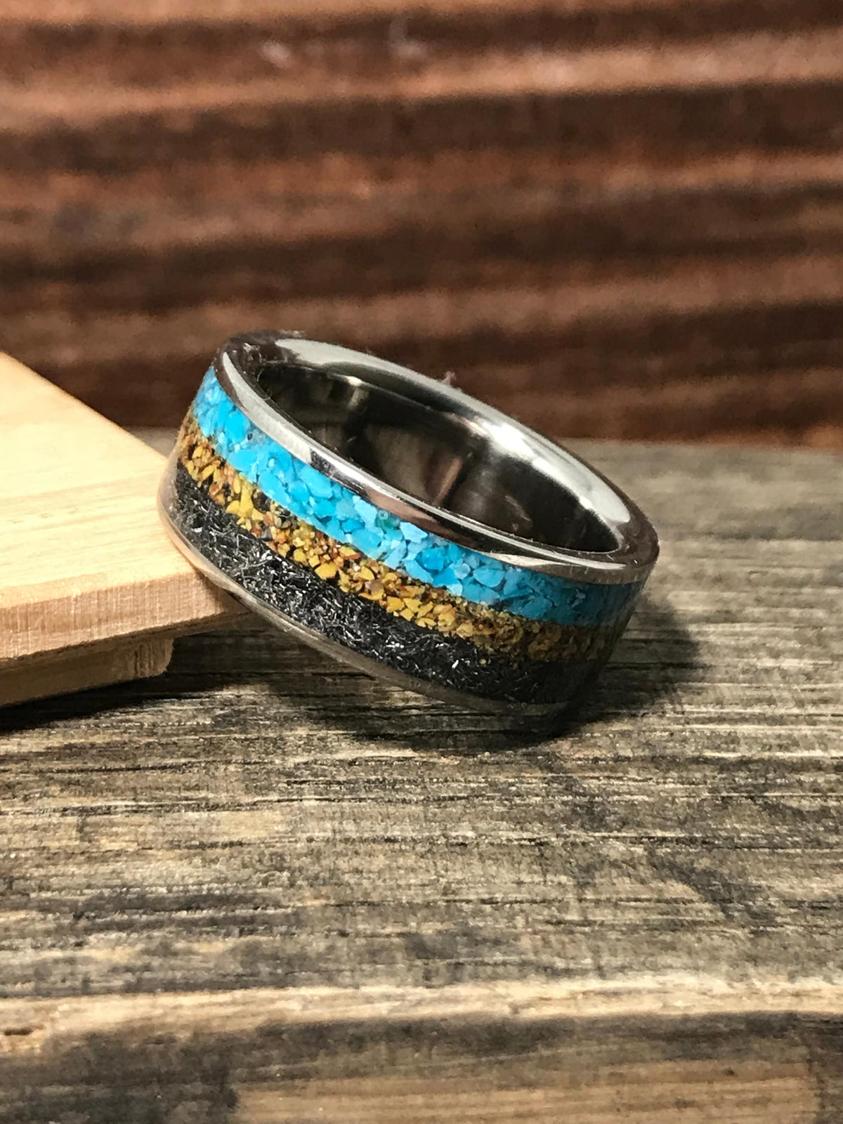 A metal band featuring three inlay stripes of bright turquoise, tan flakes, and dark textured meteorite sits angled on a wooden plank.