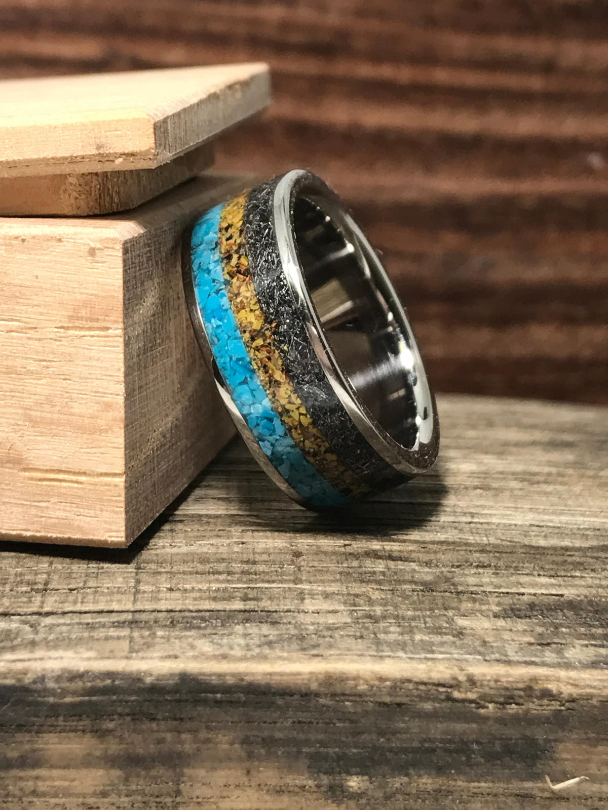 The ring is angled against the corner of a small open wooden box, displaying its turquoise, gold, and dark meteorite inlays.