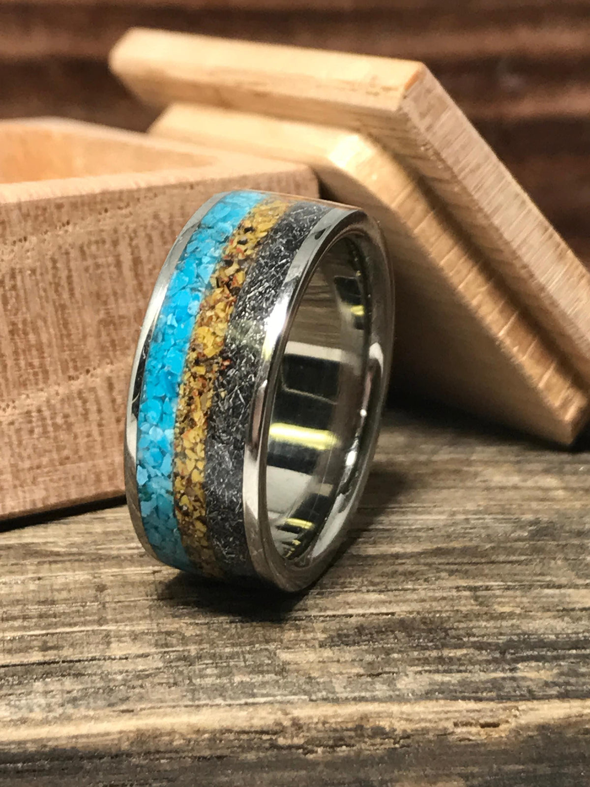 The ring stands upright on a wooden table with a small wooden box in the background, showcasing all three inlay textures.