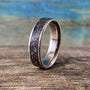 A titanium ring with a textured meteorite inlay stands upright on a rustic wooden surface against a blurred blue and brown background.