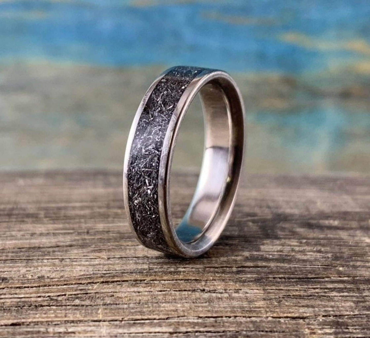 A titanium ring with a textured meteorite inlay stands upright on a rustic wooden surface against a blurred blue and brown background.