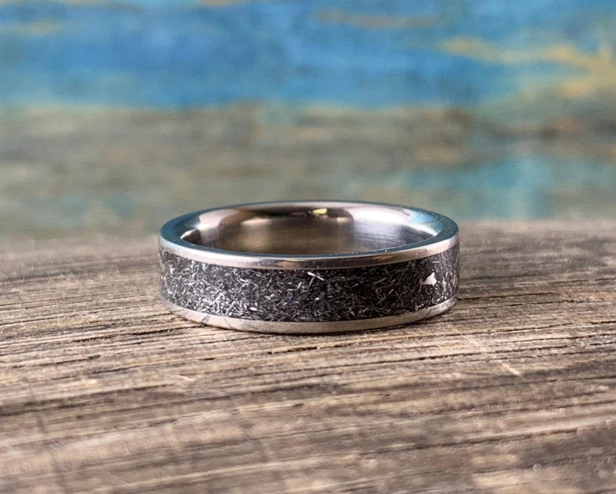 The same titanium ring lies flat on its side on a wooden board, showcasing its wide band and dark inlaid pattern.