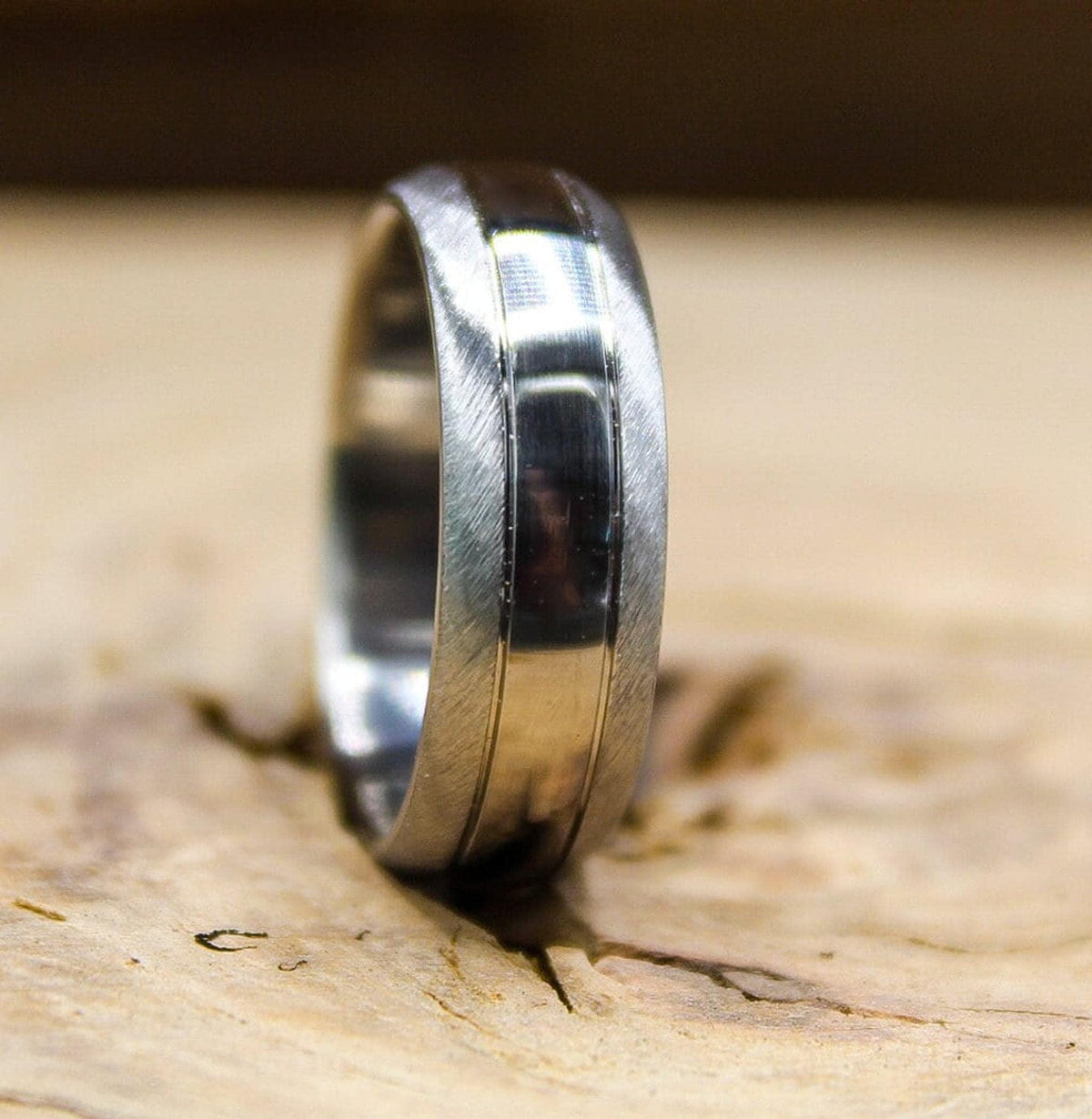A two-toned silver band with brushed edges and a shiny central channel standing upright on a textured wood block.