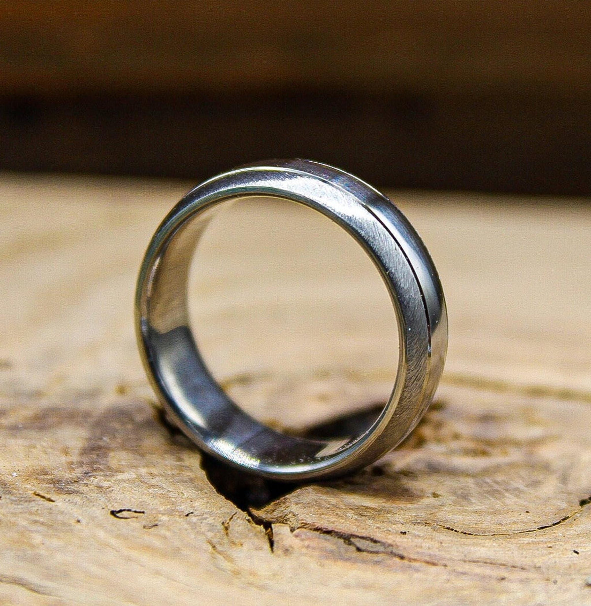 A silver metal ring with a brushed finish and a narrow polished groove angled upright on a piece of wood.