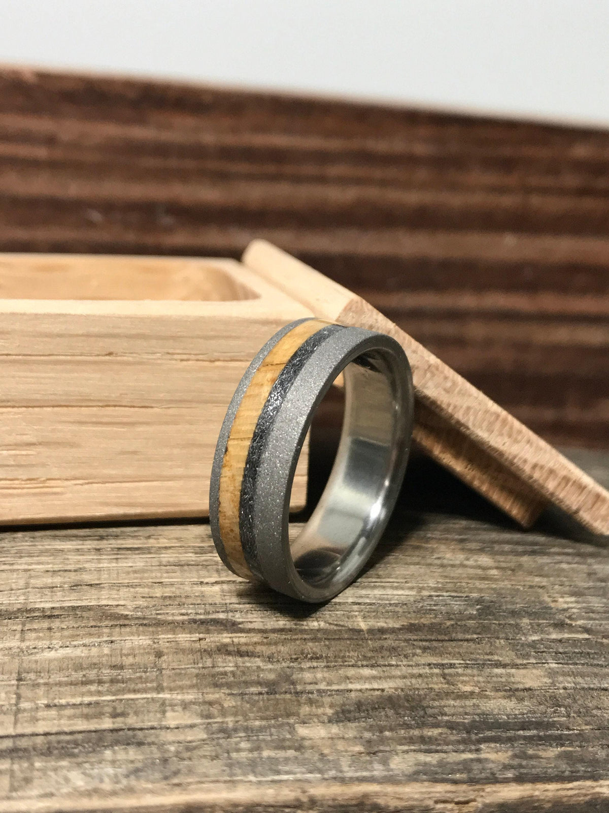 A titanium ring with wooden and dark meteorite inlays stands upright on a rustic wooden surface next to an open wooden box.