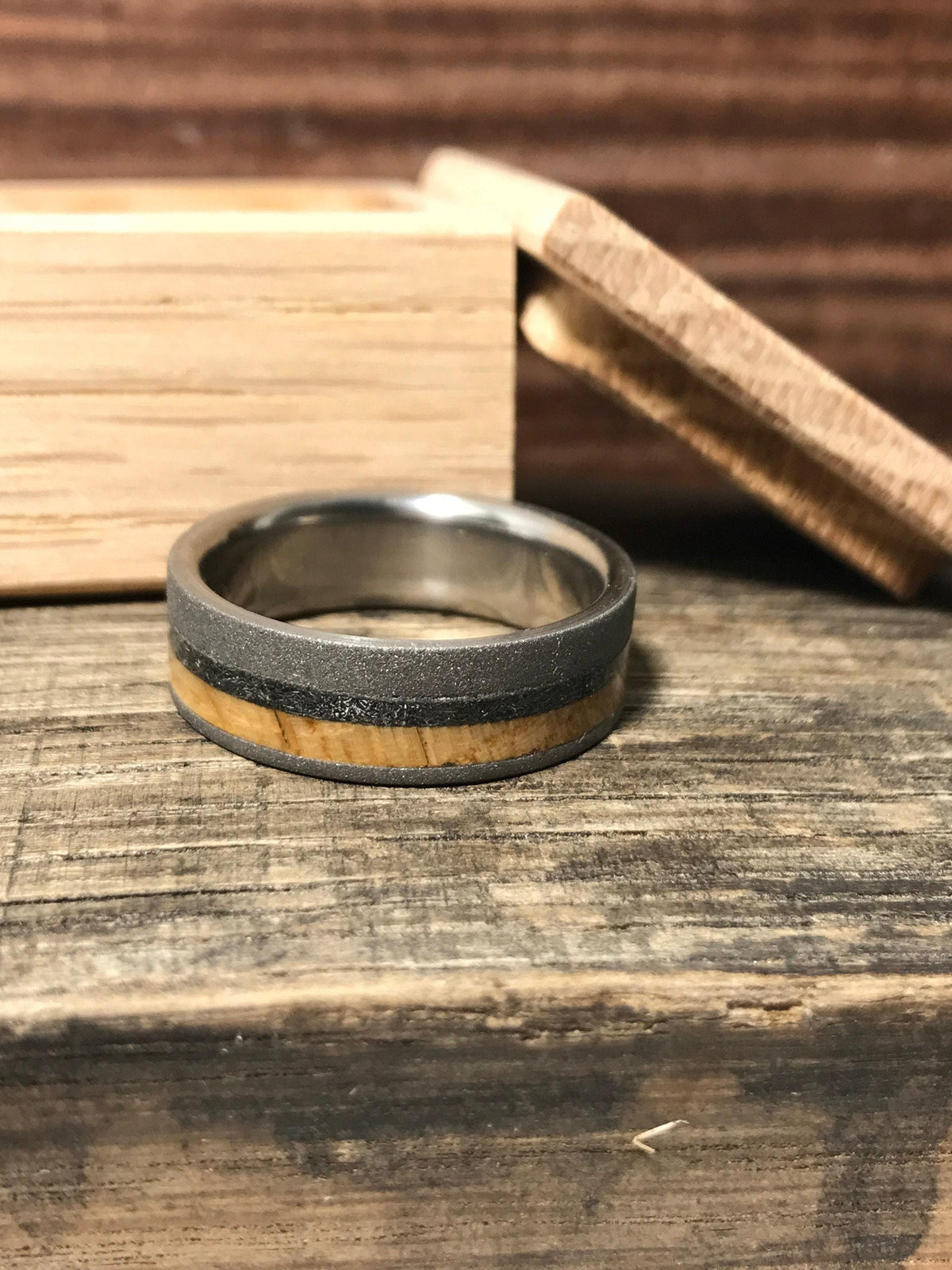 The ring with wood and meteorite inlays is displayed on a rustic wooden surface in front of a small wooden box.