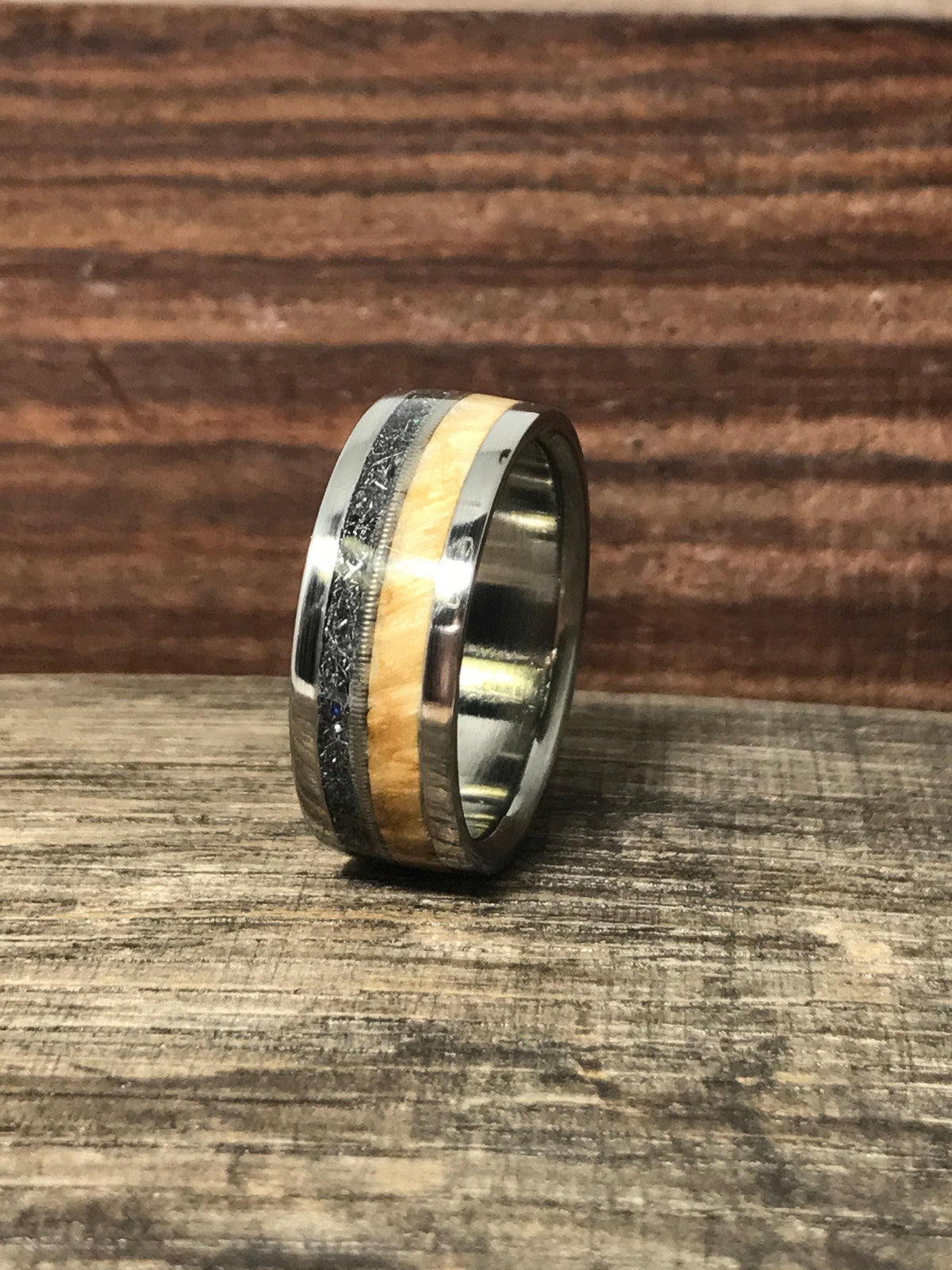 A ring featuring a metal edge, light wood inlay, guitar string detail, and textured meteorite standing upright on a wooden surface.