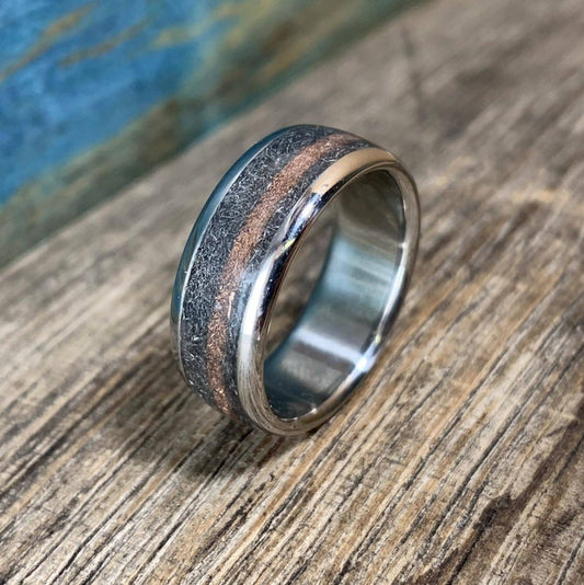 A titanium ring featuring an inlay of textured meteorite and a thin central stripe of rose gold, resting upright on a rustic wooden surface.