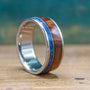 A titanium ring featuring wood and blue opal inlay rests on a wooden surface against a teal background.