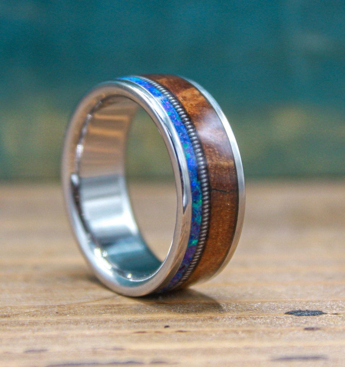 A titanium ring featuring wood and blue opal inlay rests on a wooden surface against a teal background.
