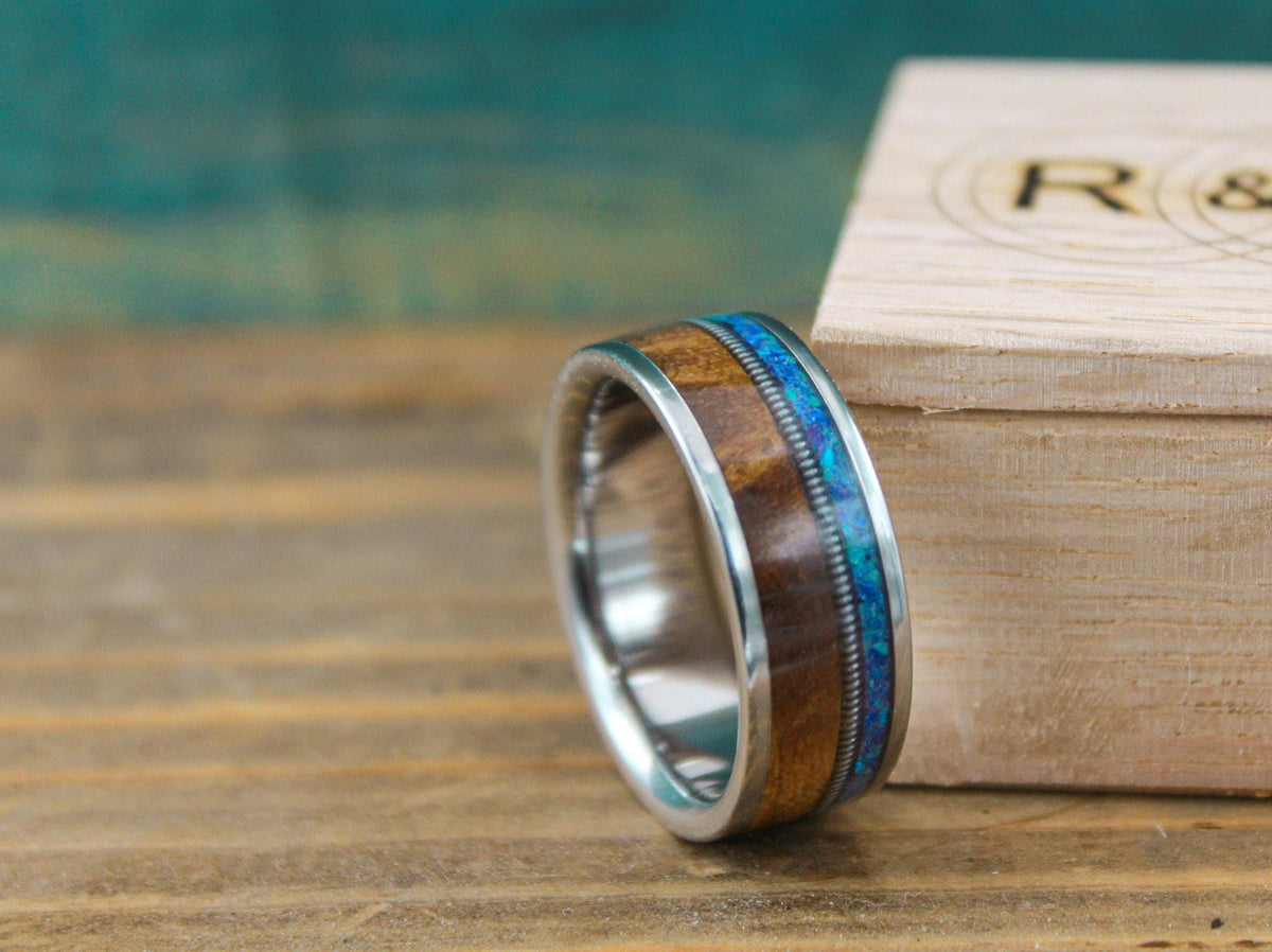 A titanium wedding band with wood and blue opal inlay is positioned beside a small wooden box.