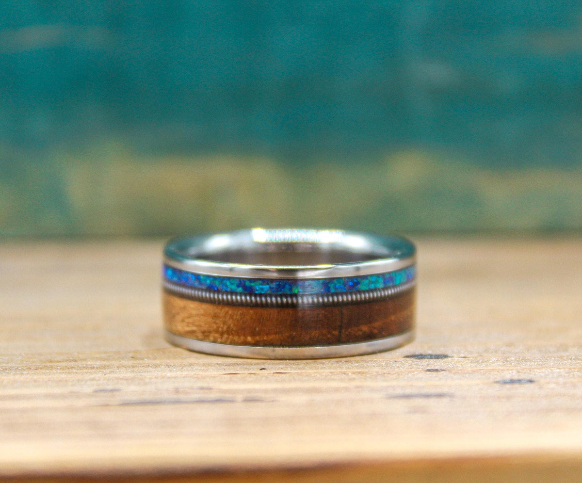 A titanium band inlaid with wood, a textured metal strip, and blue opal rests horizontally on wood.