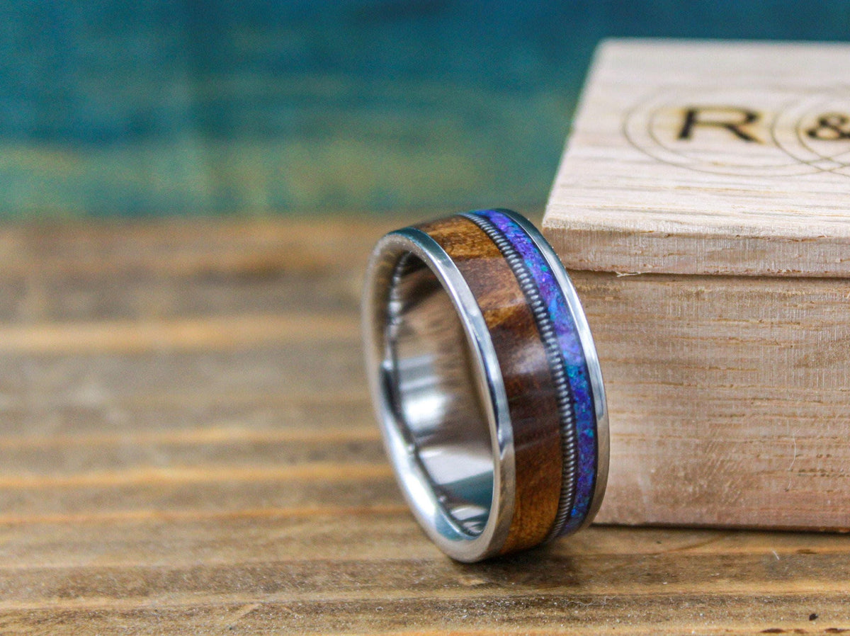 A titanium ring with wood and purple opal inlay sits in front of an engraved wooden box.