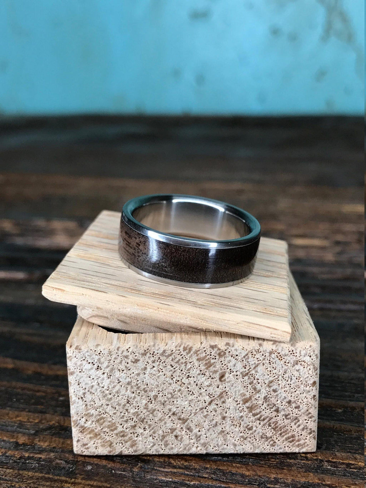 A ring with a dark wood inlay rests flat atop the slightly open lid of a small wooden box on a rustic wooden table.