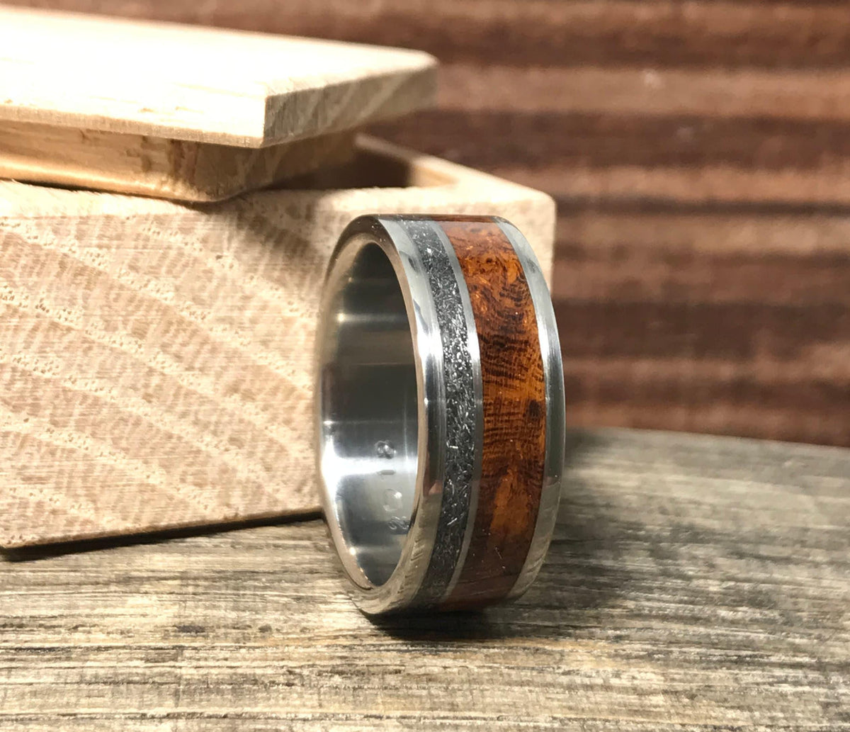 A titanium ring with meteorite and wood inlays stands upright beside a small wooden box.