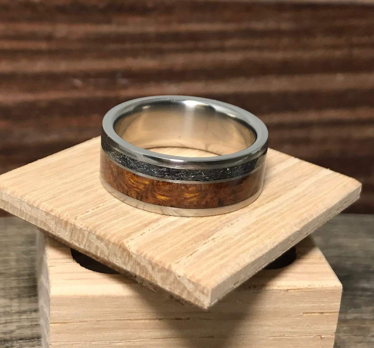 A close-up of a titanium ring with a wood and meteorite inlay sitting on a square wooden platform.