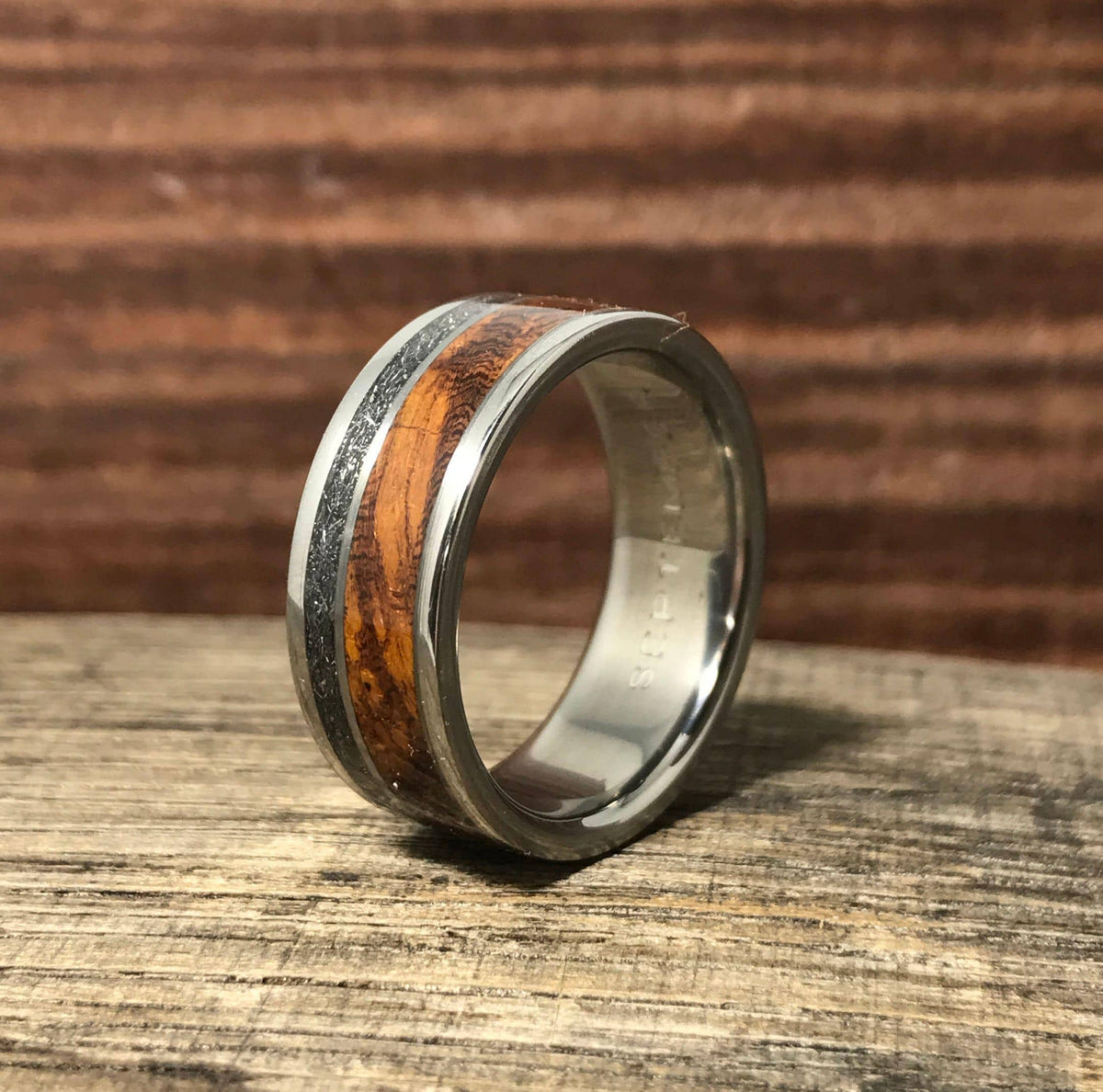 A titanium ring with meteorite and wood inlays angled upward on a rustic wooden board.