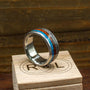 A metal ring featuring inlays of blue opal, red wood, and metallic meteorite rests at an angle on a small wooden box stamped with the letters R and L.