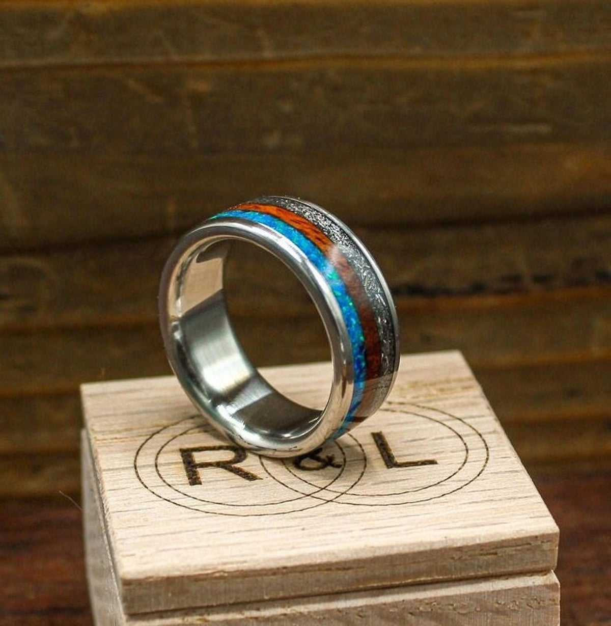 A metal ring featuring inlays of blue opal, red wood, and metallic meteorite rests at an angle on a small wooden box stamped with the letters R and L.