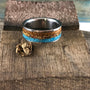 A metal ring with wood, stone, and bone inlays rests on a rustic wooden block with a small wood fragment beside it.