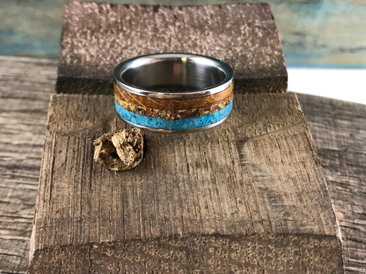 A metal ring with wood, stone, and bone inlays rests on a rustic wooden block with a small wood fragment beside it.