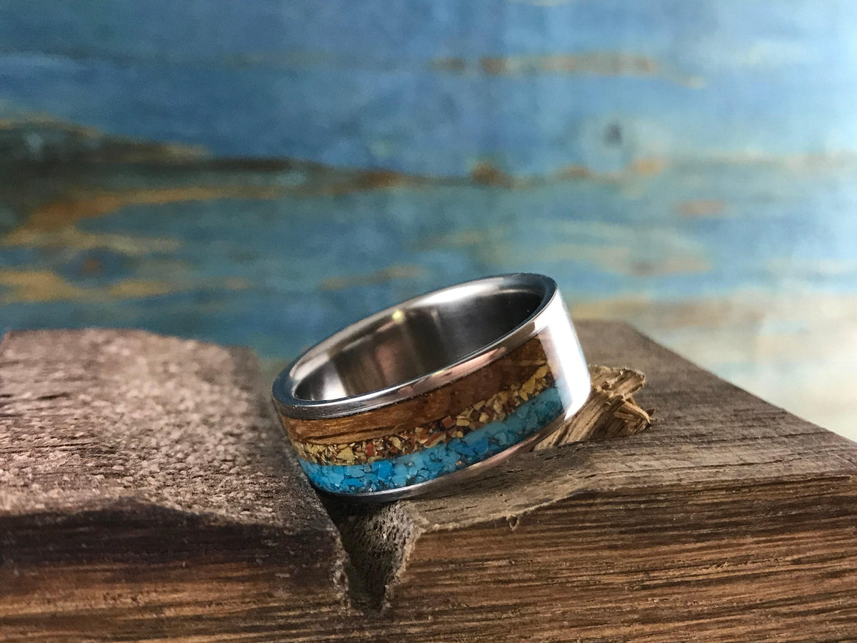 A ring featuring inlaid wood and turquoise sits upright on a piece of weathered wood against a blue and brown background.