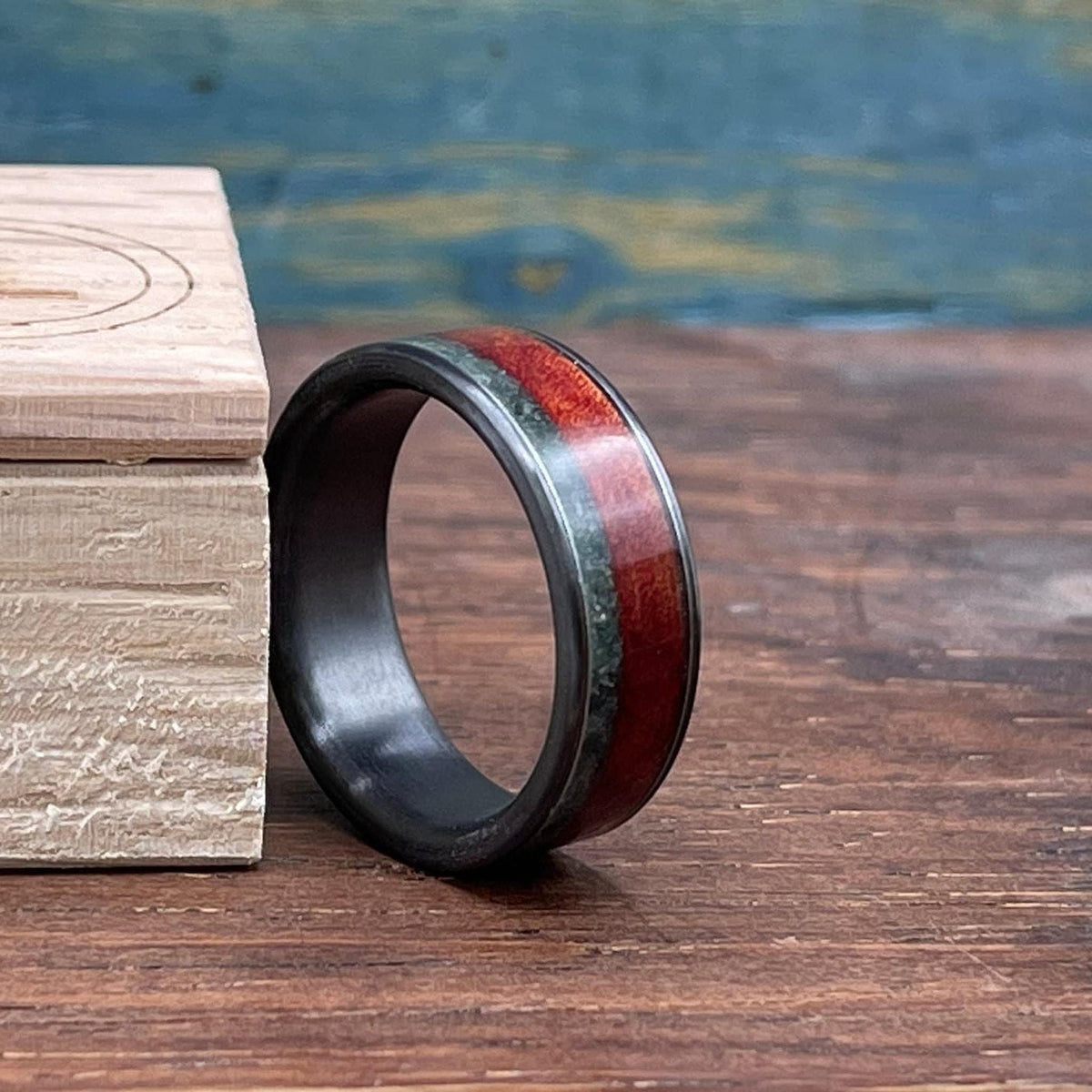 The ring featuring red wood and green stone inlays sits upright next to a small wooden box against a textured blue and brown backdrop.