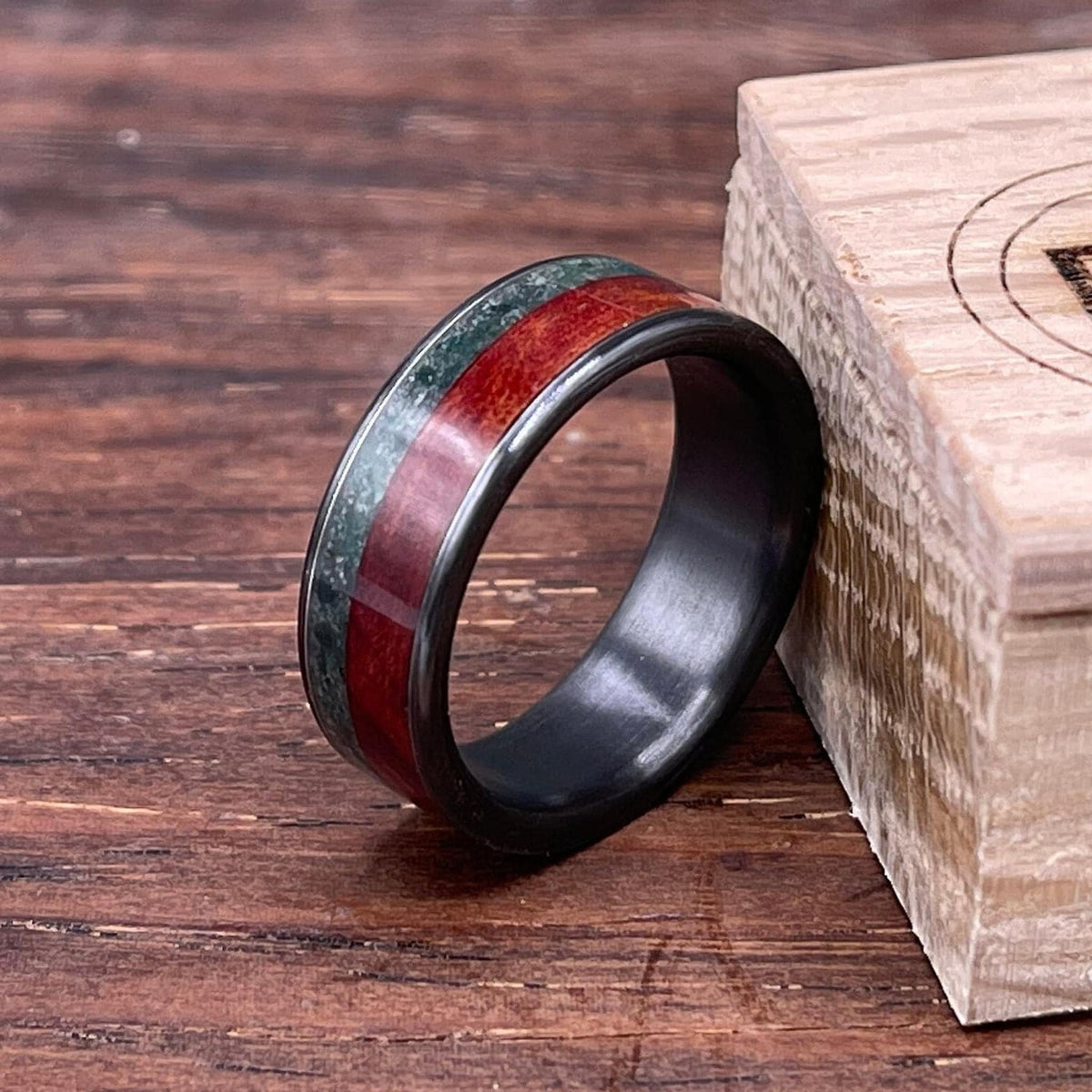 An angled view of the black ring with red and green inlays positioned beside the corner of a stamped wooden box on a dark wooden table.