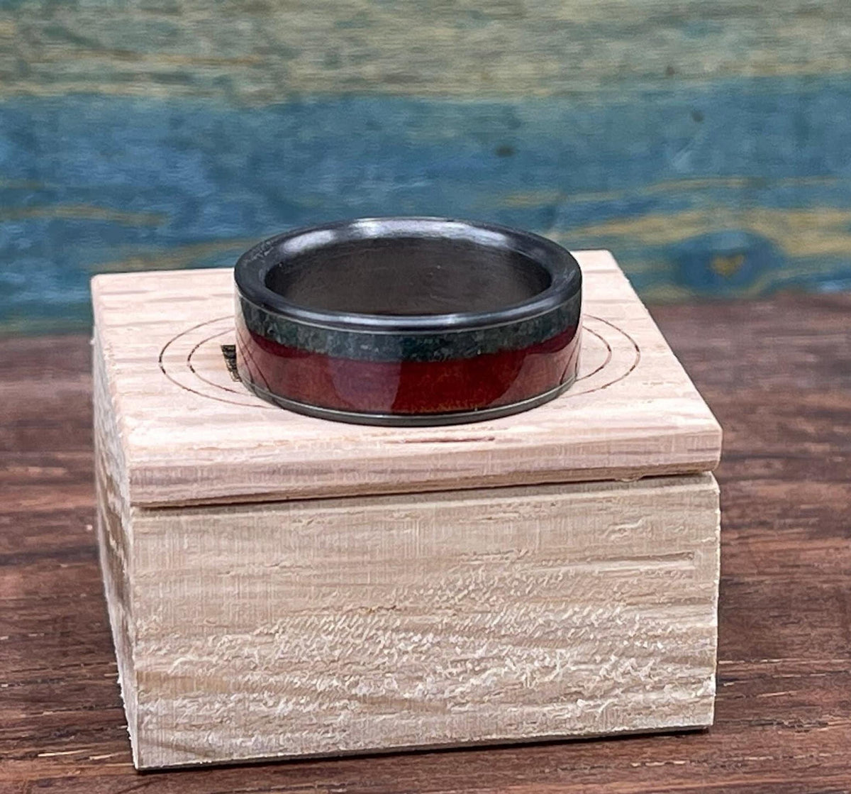 The ring rests flat on top of a small square wooden box with circular branding on its lid.