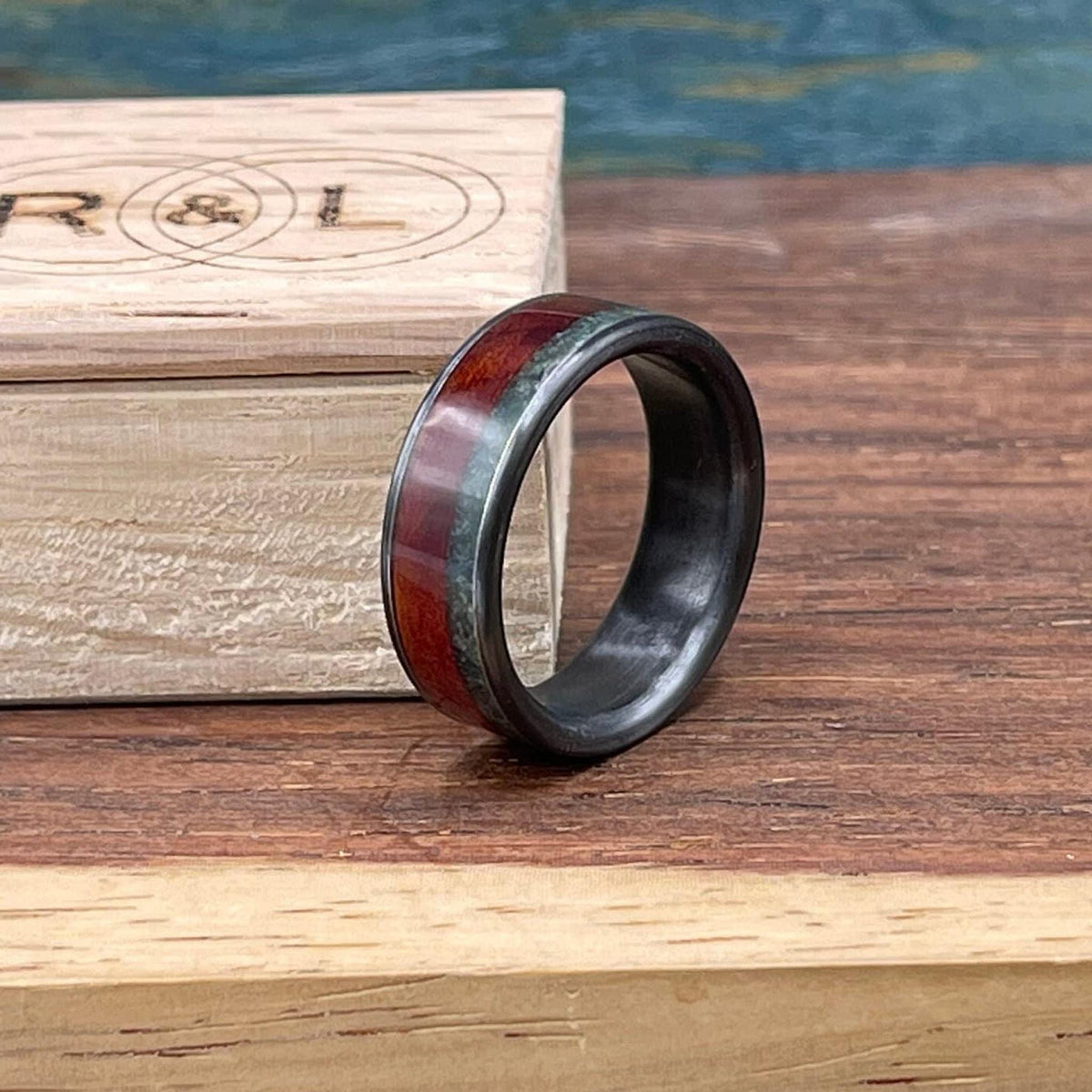 The ring stands upright on a wooden surface with an engraved wooden box bearing initials R and L visible in the background.