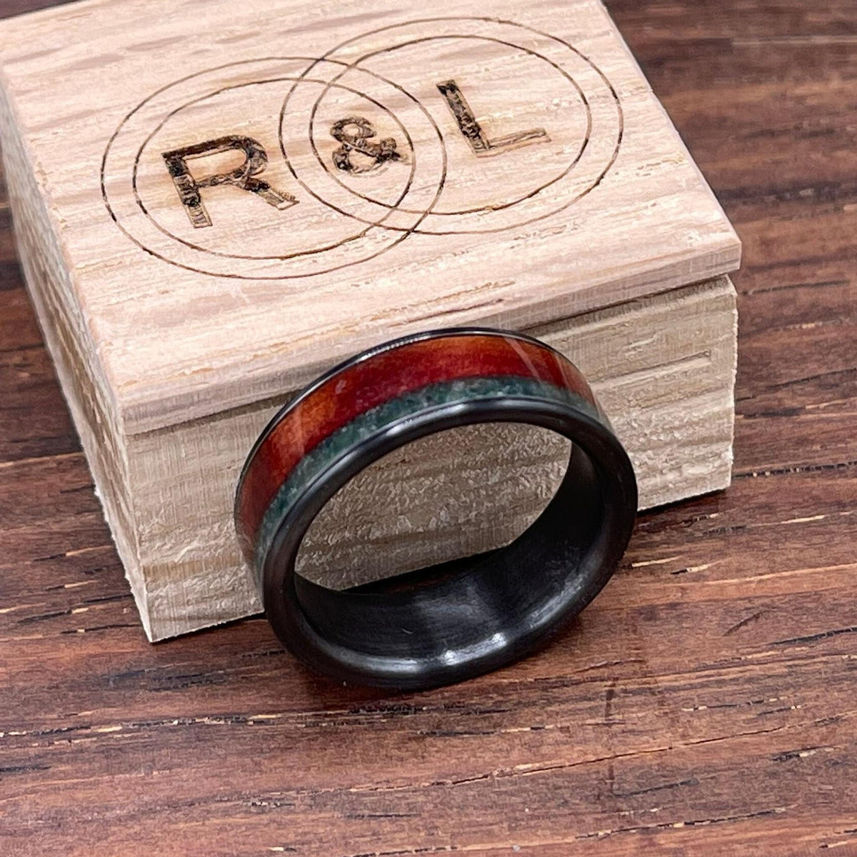 The black ring with red wood and green stone inlays rests in front of a branded wooden box top on a wood-grain surface.