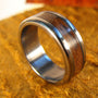 A silver metal ring with a wide wood grain inlay resting on a textured gold-colored surface.