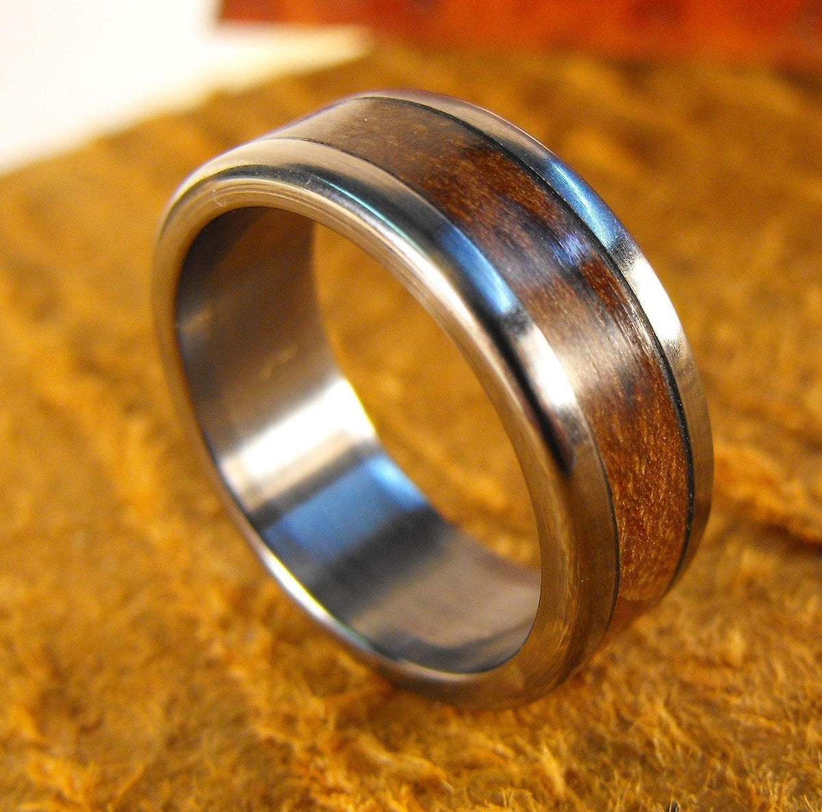 A silver metal ring with a wide wood grain inlay resting on a textured gold-colored surface.