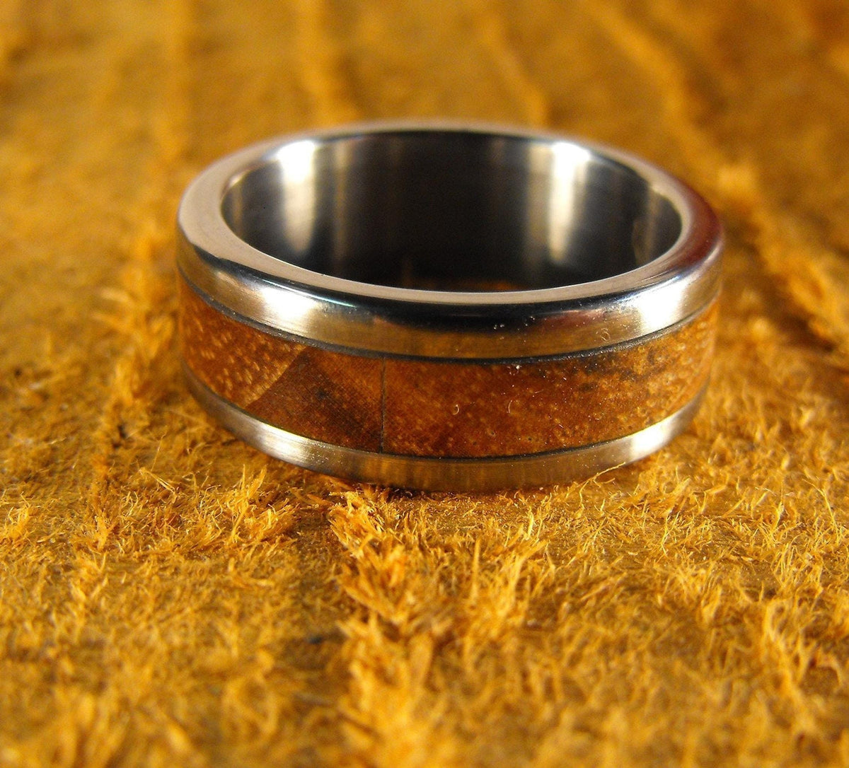 A metal wedding band featuring a central wood inlay, viewed from a slightly high angle on a golden textured background.