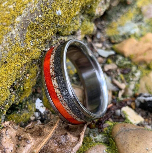 A titanium ring featuring an inlay of dinosaur bone, meteorite, and wood, resting upright on mossy stone outdoors.