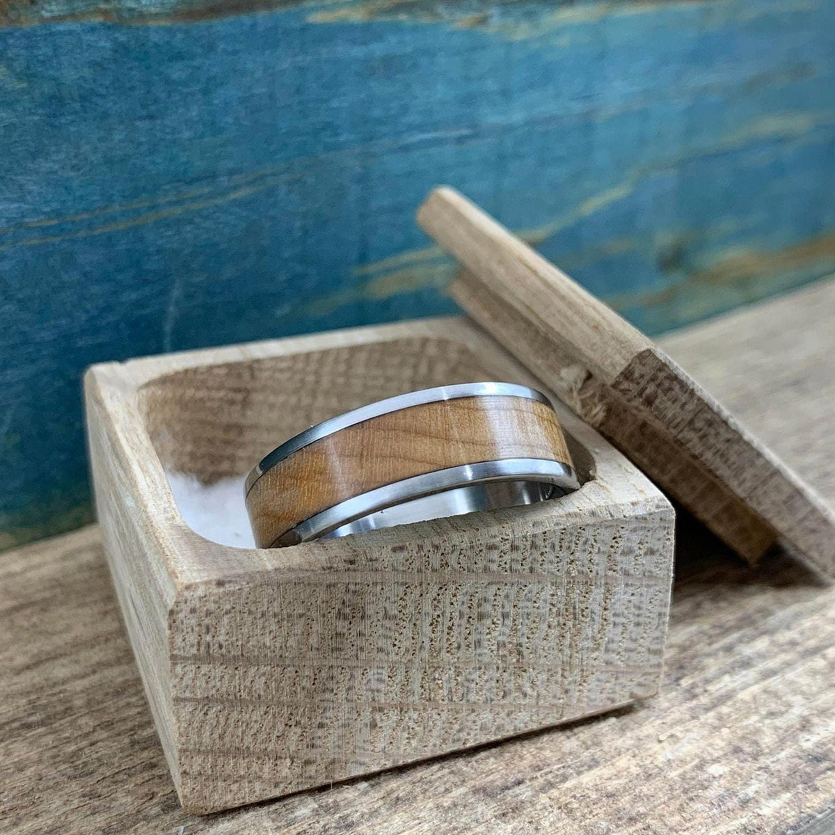 A metal ring with a wood inlay sits inside a small wooden box on a textured wooden surface against a blue backdrop.