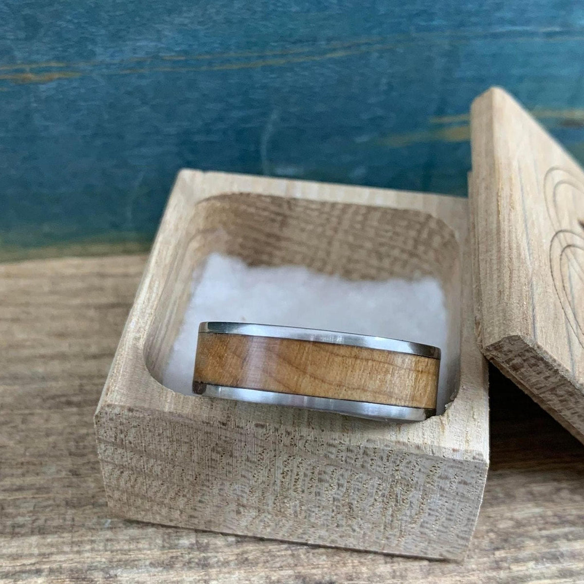 A ring with a wooden inlay rests in a small open wooden box filled with white shredded paper packaging.
