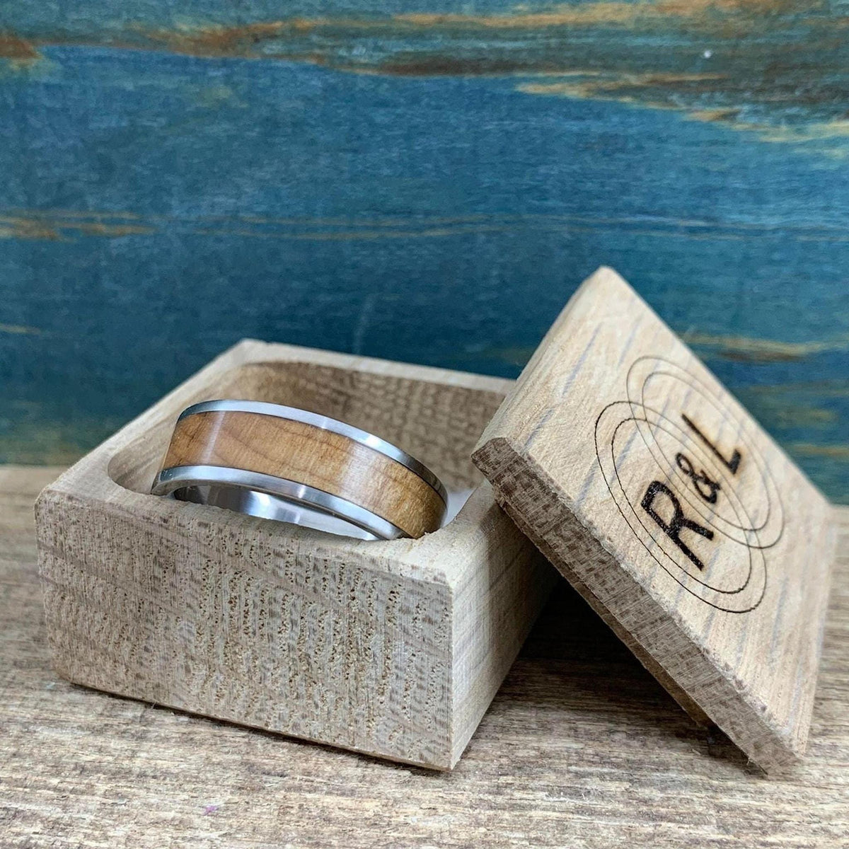 A metal ring with a wood inlay is placed in a small wooden box, with the box lid leaning open showing engraved letters.