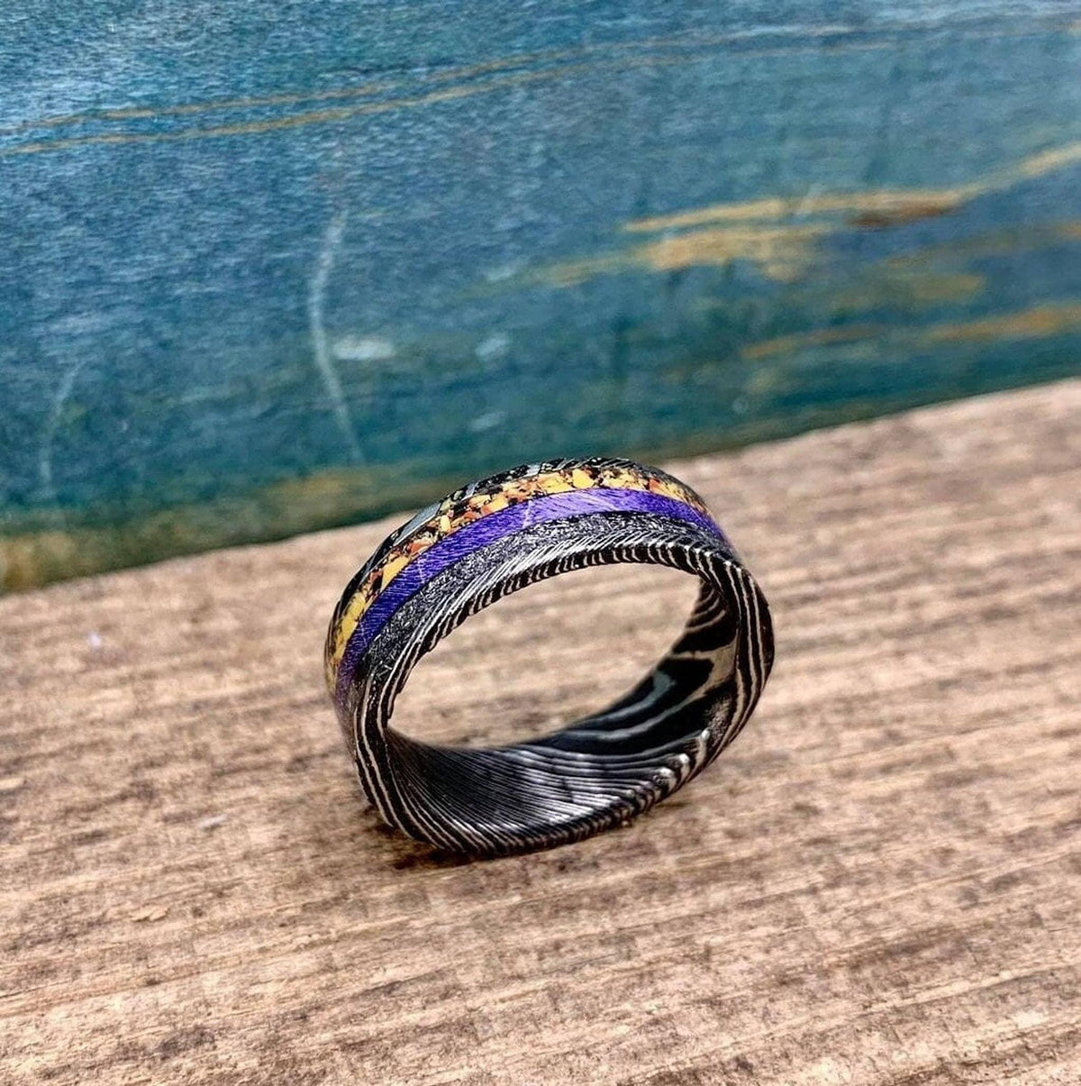 A patterned Damascus steel ring with purple and textured inlays resting on a rustic wooden surface.