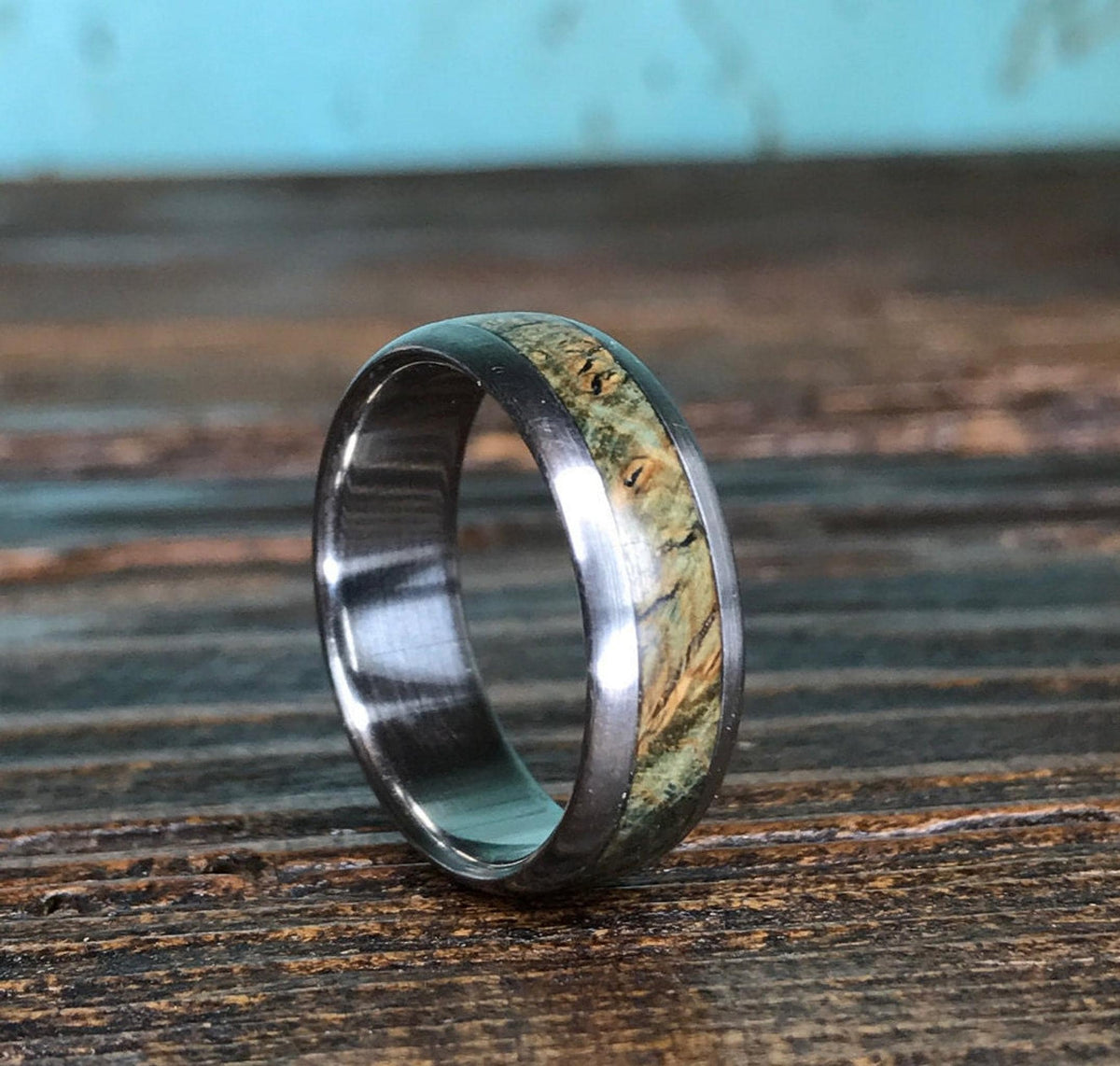 A titanium ring with a textured wood inlay sits angled on a weathered wooden plank against a light blue background.