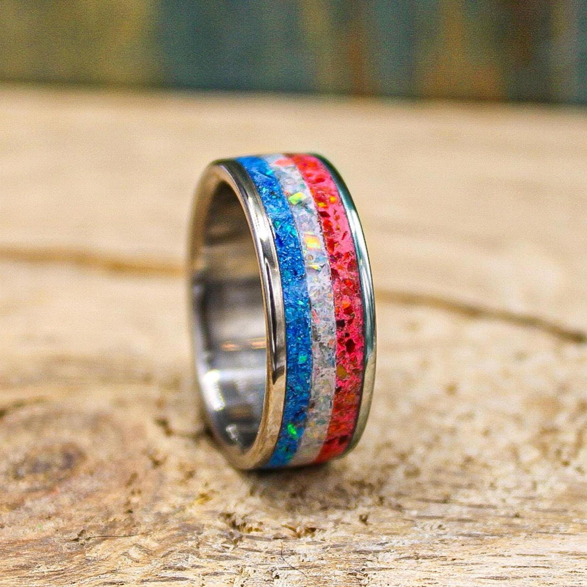 The same titanium ring shown at a slightly different angle on the wood grain surface, displaying its blue, white, and red inlaid channels.