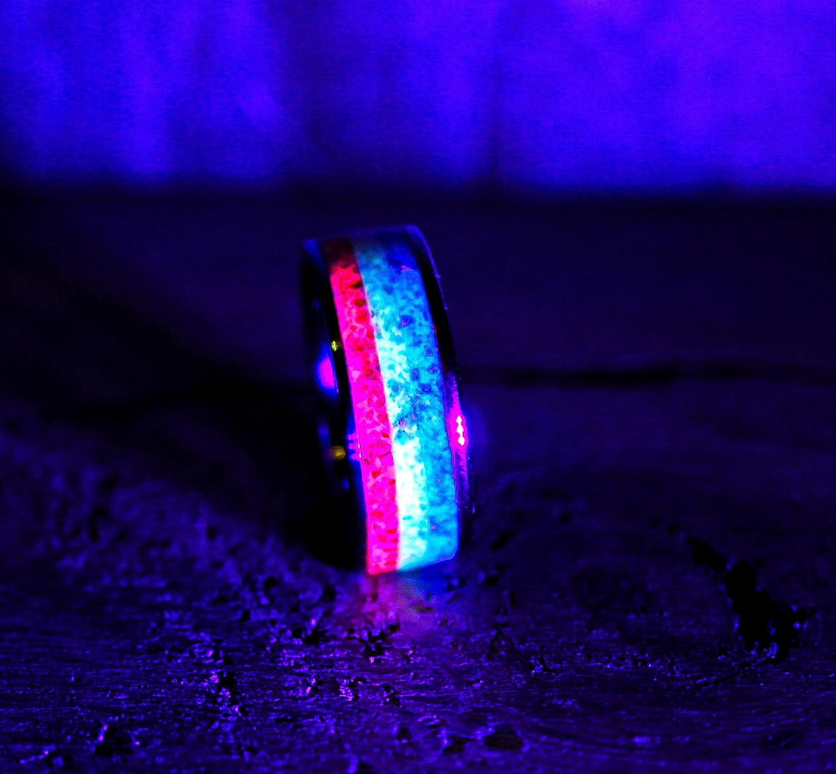 The ring glowing in the dark under UV light, casting vibrant pinkish-red and bright cyan light from its inlaid channels against a dark background.