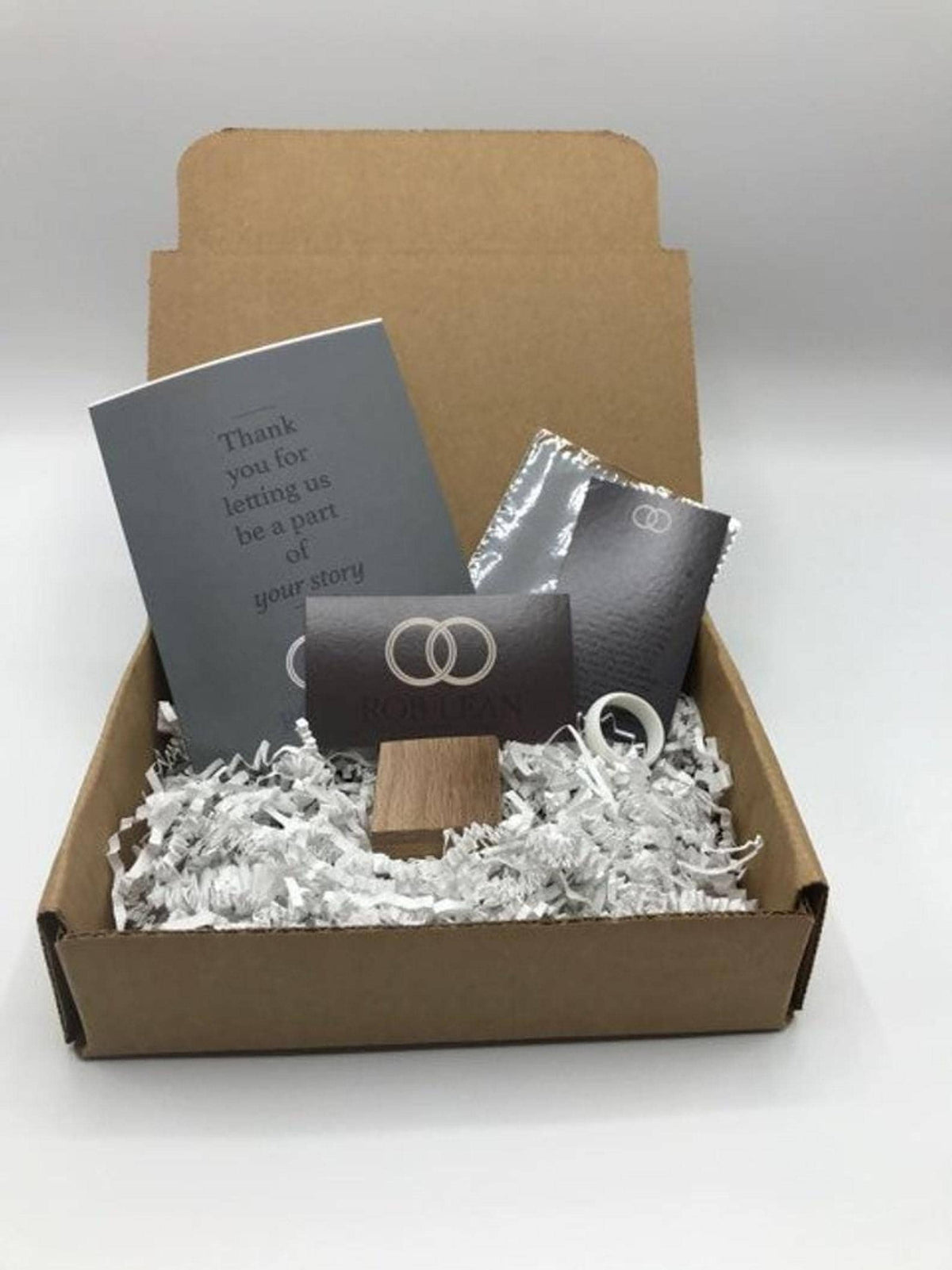 An open cardboard shipping box filled with white shredded paper packaging, containing a grey greeting card, a small wooden ring box, a white ring, and branding cards.